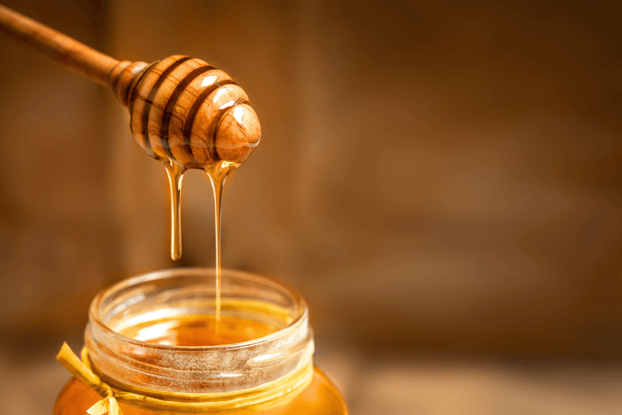 A wooden honey dipper drips honey into a jar against a blurred background.