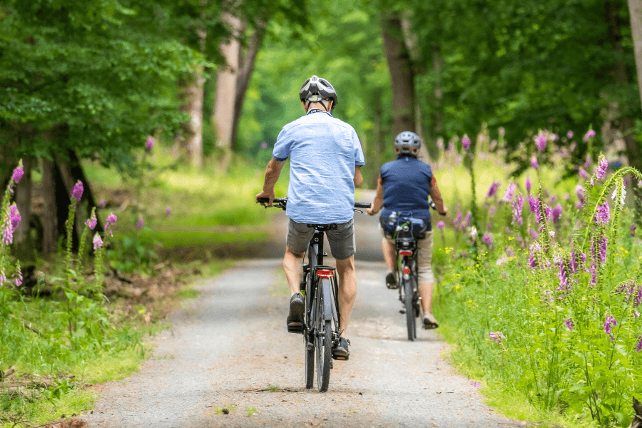 Two cyclists ride on a gravel path surrounded by green trees and wildflowers.