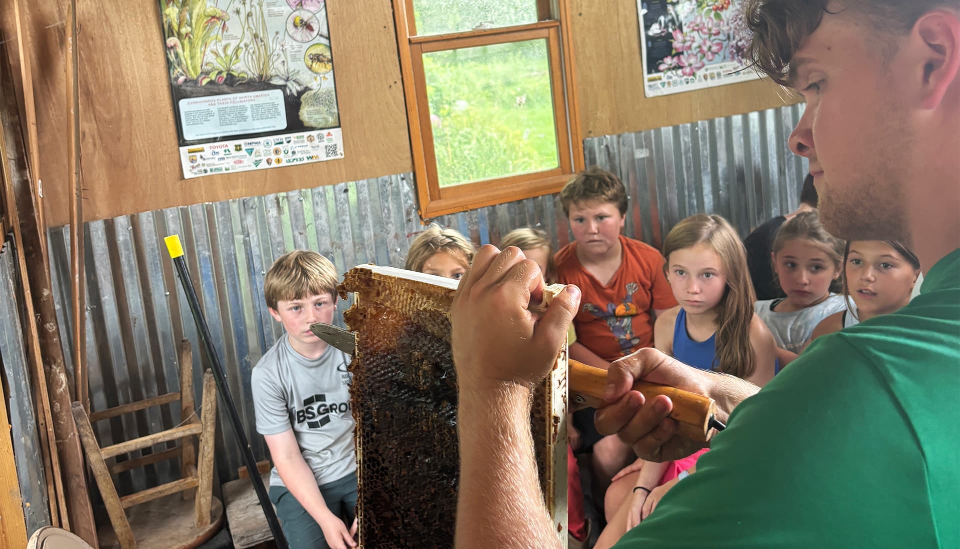 A group of children watches intently as a young man demonstrates beekeeping with a honeycomb frame.