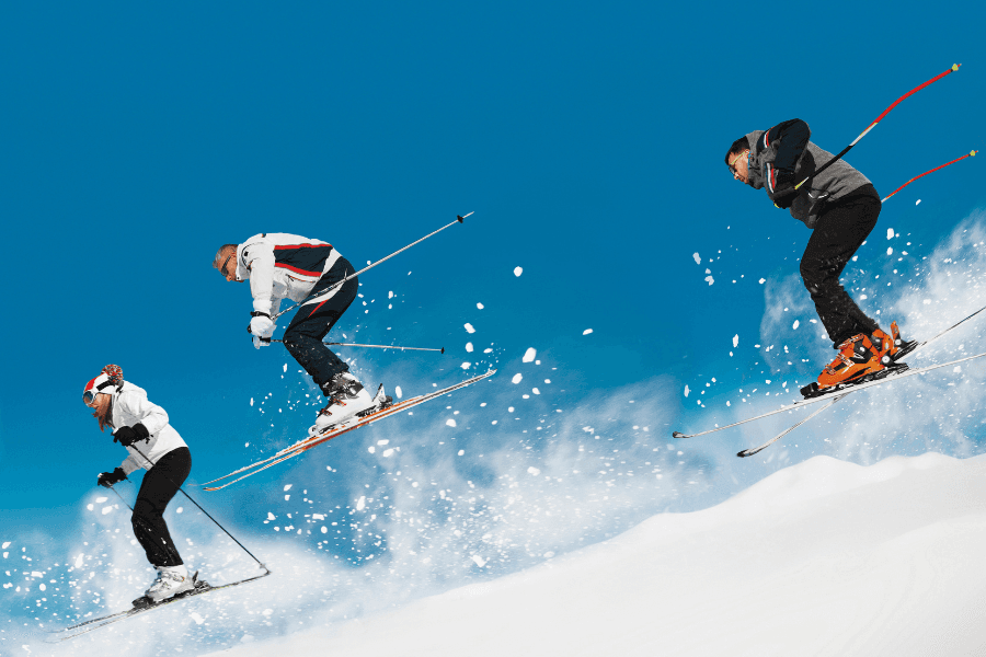 Three skiers jump through the air against a clear blue sky, creating snow spray below.