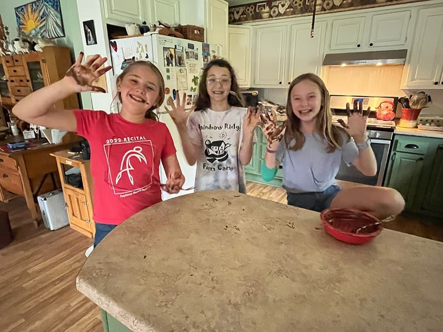 Three smiling girls show their chocolate-covered hands in a kitchen.
