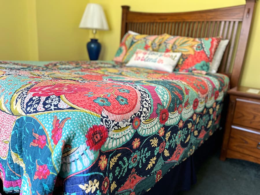 A vibrant, patterned quilt covers a bed in a brightly colored room with a lamp and nightstand nearby.