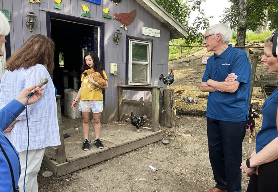 A young girl holding a chick speaks to a group of adults outside a barn, with chickens roaming the yard.