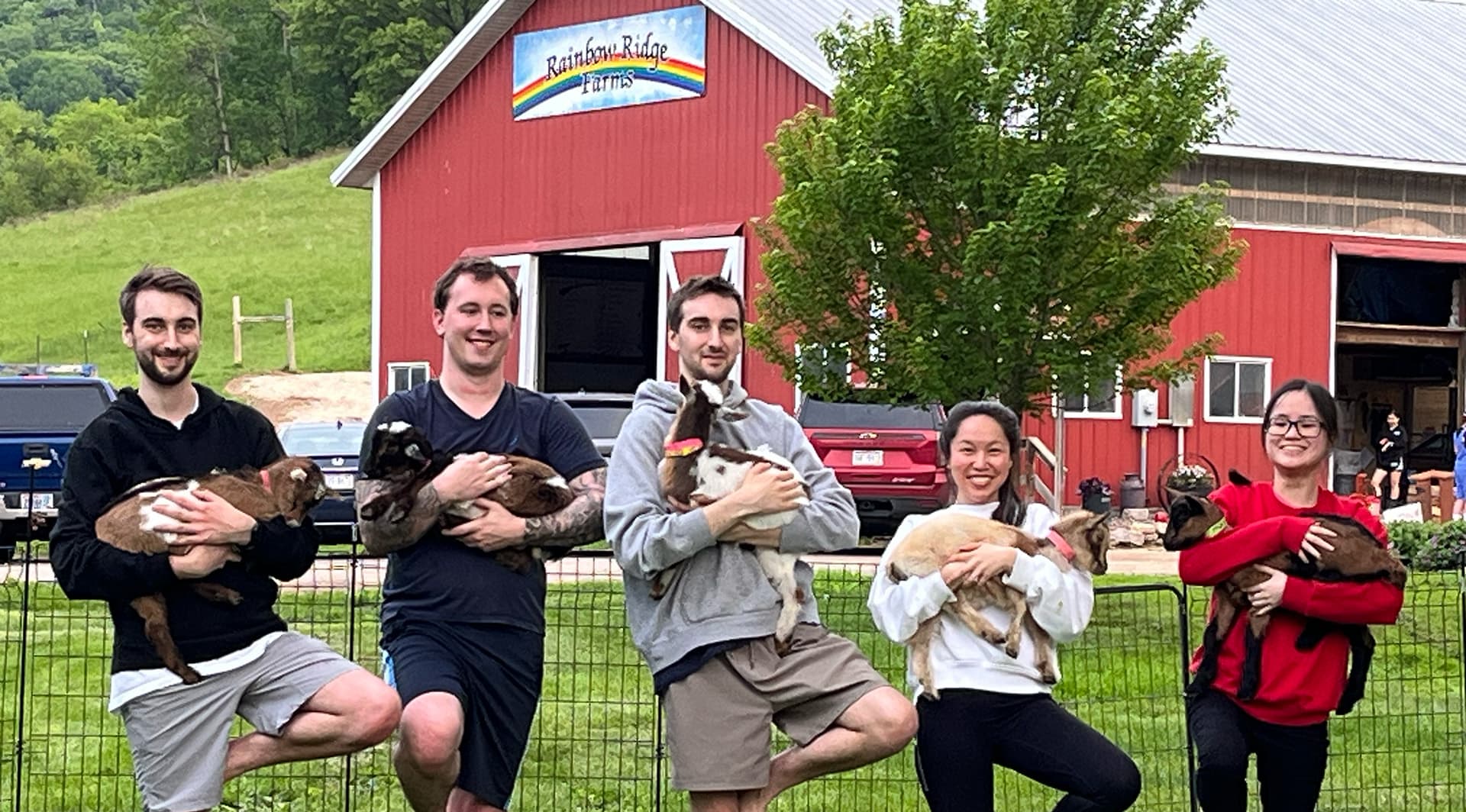A group of five people standing in a grassy area, each holding a goat in front of a red barn.