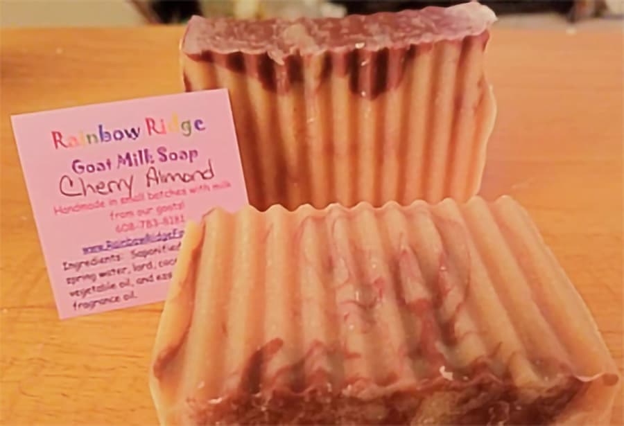 A close-up of two bars of Cherry Almond goat milk soap with a colorful informational card beside them.