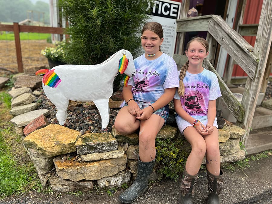 Two girls in tie-dye shirts sit on a stone wall next to a colorful, painted goat statue.