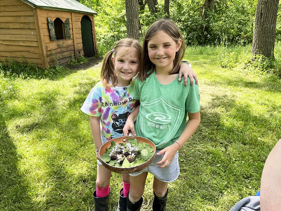 Two young girls stand in a grassy area, holding a bowl of greenery and flowers, smiling together.