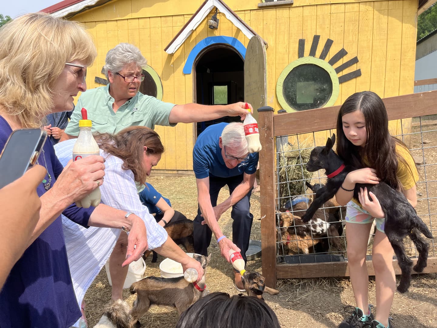 A group of people interacts with baby goats at a farm, with one child holding a goat while others feed them.
